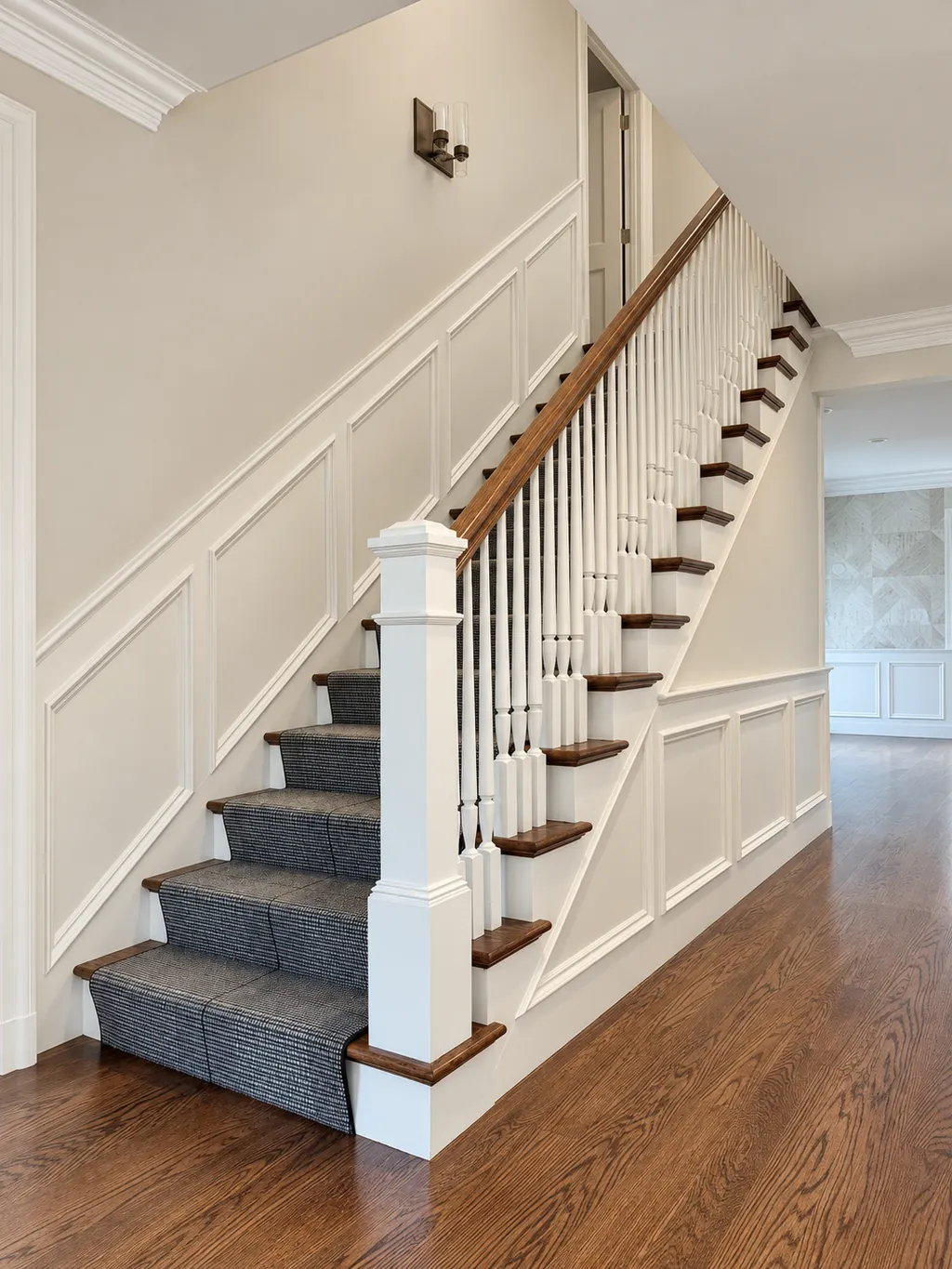 Straight staircase with a white paneled newel post, turned balusters and a stained oak handrail, rising along raised panel wainscoting